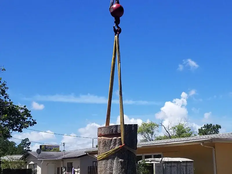 A large cut trunk section hanging in nylon slings from a crane hook, lifted clear above the rooflines of a residential street.