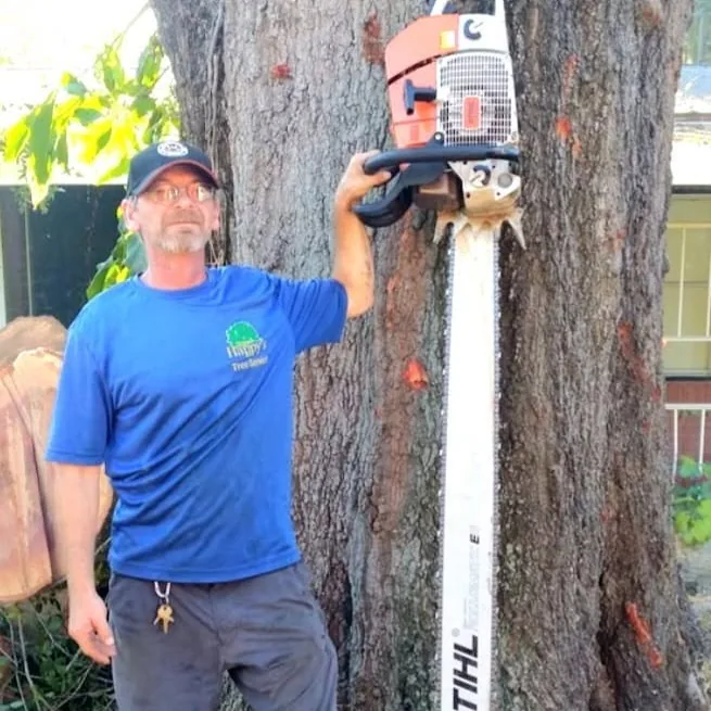Craig, a Happy's Tree Service crew member in a branded blue shirt, standing against a large tree trunk and holding a chainsaw whose guide bar is nearly as tall as he is.