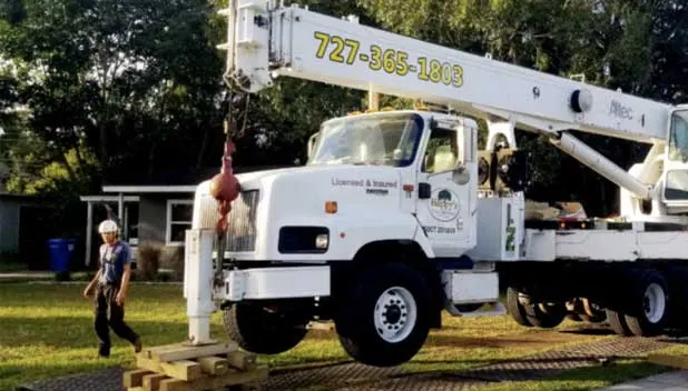 A boom truck with a hook block, set up on outrigger mats, the phone number painted along the boom and Licensed and Insured lettering on the door, a crew member in a white helmet walking past.