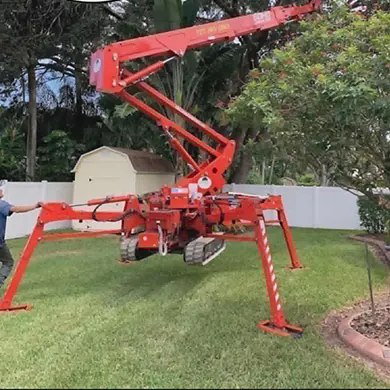 An orange tracked spider lift standing on a back lawn with all four outrigger legs down on the grass and its articulated boom raised but still folded at the knuckle, a crew member in a cap setting one of the outriggers by hand, a white vinyl privacy fence and a garden shed behind.